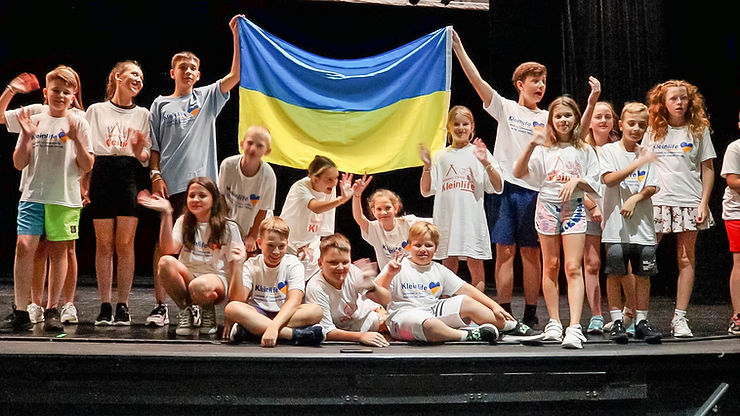 Ukrainian Refugee Summer Camp campers in KleinLife's theater holding up Ukrainian flag and waving to the camera.