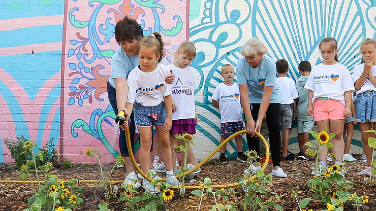 Victoria Faykin embraces one camper outside in a garden at KleinLife. They both are holding a hose together to water a garden bed of sunflowers. A teacher helps hold the hose behind them and several campers look on while standing behind them, too. They are all in front of a colorful wall mural.