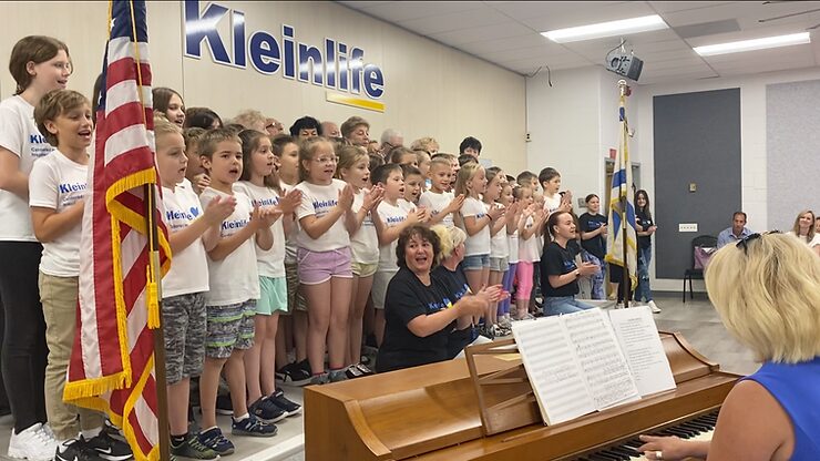 Children ages 6-12 and Holocaust survivors gather on a raised stage with the American flag on the left side of the stage and an Israeli flag on the right side of the stage are singing and clapping to "God Bless America" while it is being played on the piano. They are wearing white t-shirts with the KleinLife logo on them along with a heart-shaped Ukraine flag.